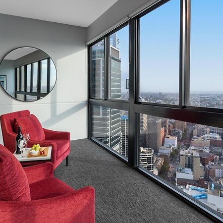 Meriton Suites Kent Street, Σίδνεϊ Εξωτερικό φωτογραφία The image shows a modern interior space with large windows that provide a panoramic view of a cityscape. In the foreground, there are two red armchairs positioned near the windows. A round mirror is mounted on the wall beside the chairs. A small tabl