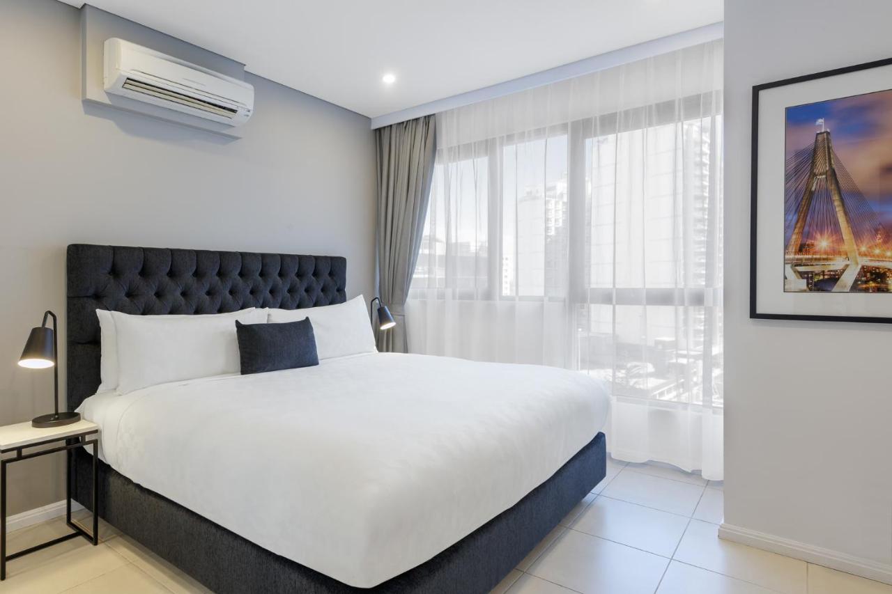 Meriton Suites Kent Street, Σίδνεϊ Εξωτερικό φωτογραφία The photo shows a modern hotel room featuring a neatly made bed with a dark, tufted headboard and white bedding. A small gray decorative pillow is placed at the center of the bed. To the left, there's a bedside table with a lamp. Large windows dresse