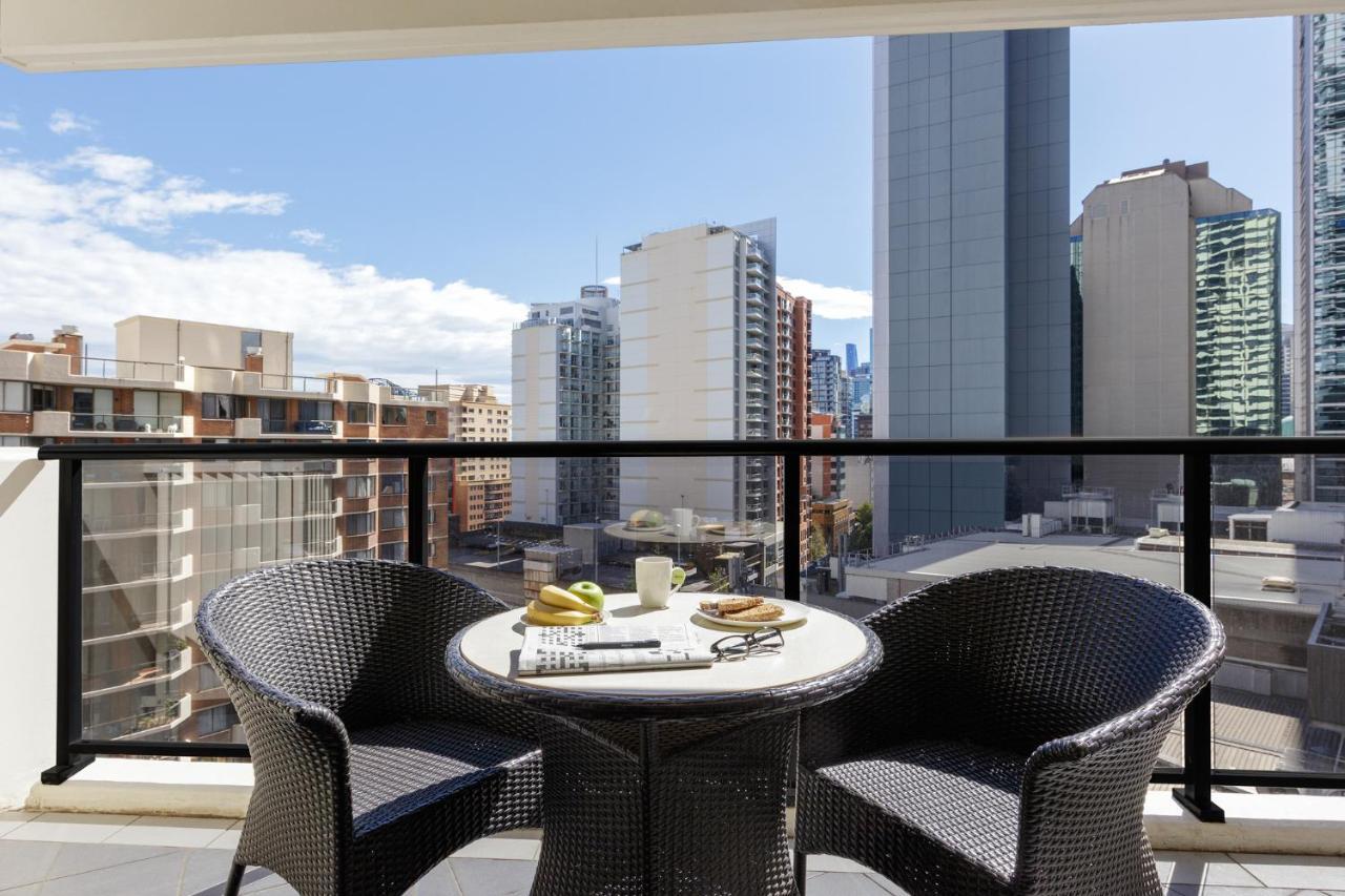 Meriton Suites Kent Street, Σίδνεϊ Εξωτερικό φωτογραφία The photo shows a balcony with a modern setting. There are two wicker chairs and a round table. On the table, there are a few items, including a cup, a plate with some snacks, and a magazine or newspaper. In the background, a cityscape can be seen, f