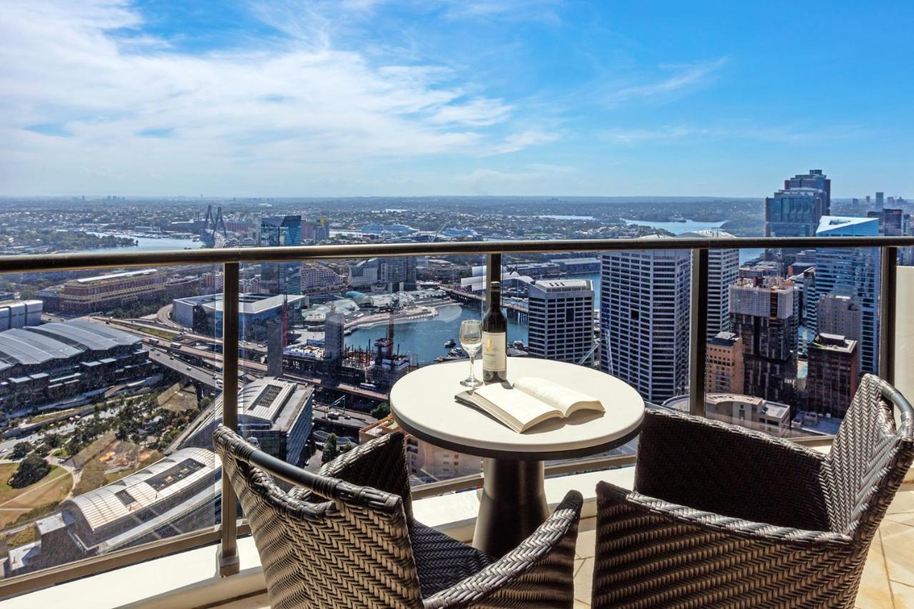 Meriton Suites Kent Street, Σίδνεϊ Εξωτερικό φωτογραφία The photo shows a beautiful panoramic view from a high balcony. In the foreground, there is a round table with a bottle of wine and an open book on it. Surrounding the table are two comfortable-looking chairs made of woven material. The background fe