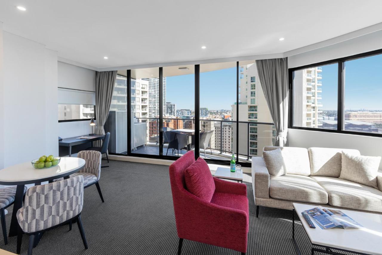 Meriton Suites Kent Street, Σίδνεϊ Εξωτερικό φωτογραφία The photo shows a modern, well-lit living space with large windows providing a view of a cityscape. There is a comfortable seating area featuring a gray sofa and a red accent chair. A small round dining table with two chairs is situated to the left, 