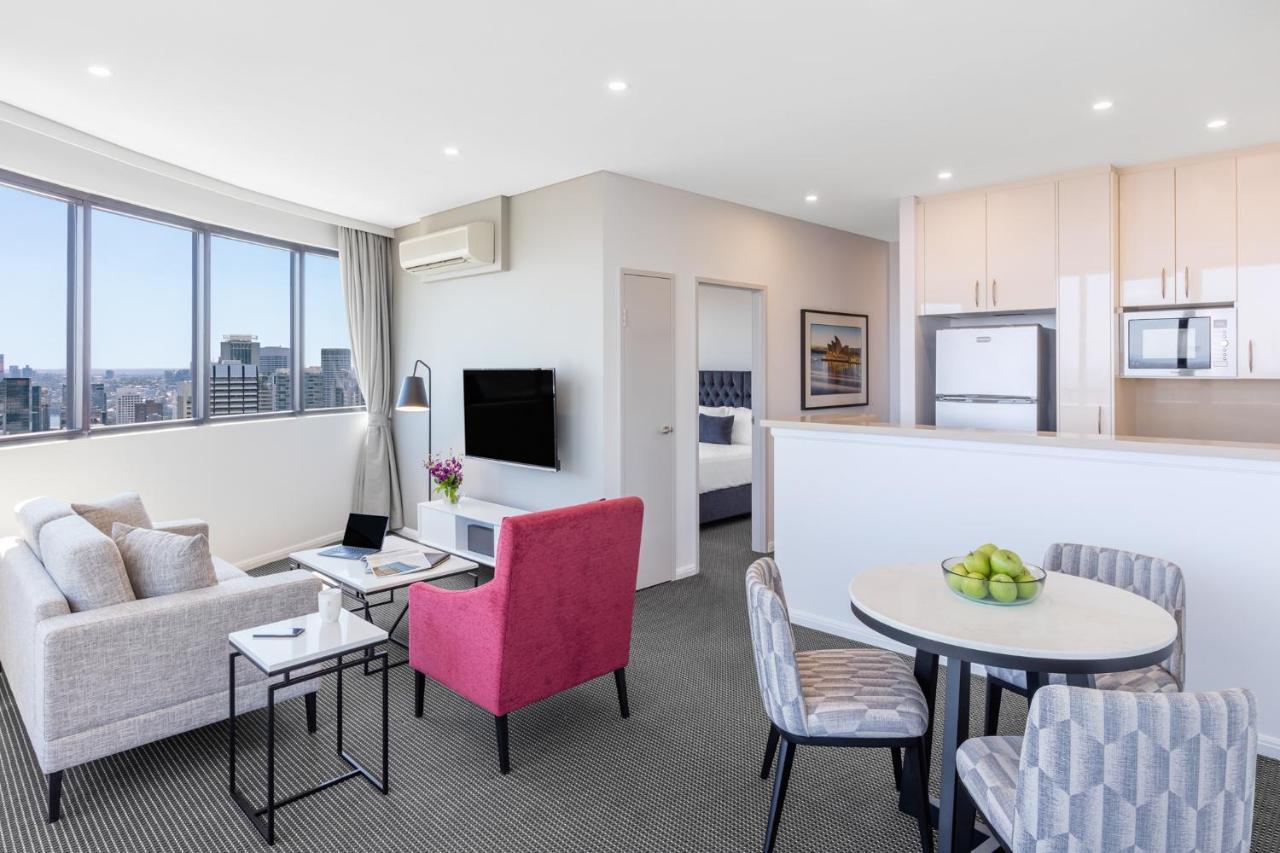 Meriton Suites Kent Street, Σίδνεϊ Εξωτερικό φωτογραφία The photo shows a modern living space, possibly a hotel room or an apartment. It features a cozy seating area with a light-colored sofa and a pink armchair. There is a small black table beside the sofa and a round dining table with gray chairs, toppe