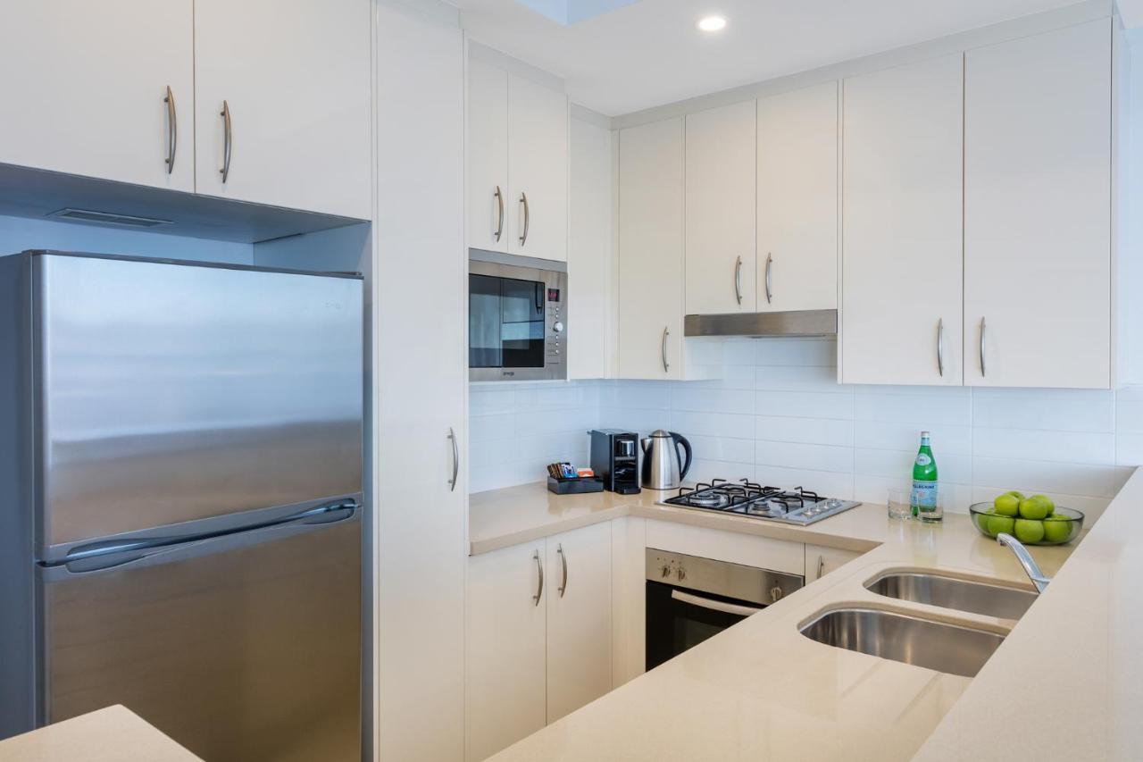 Meriton Suites Kent Street, Σίδνεϊ Εξωτερικό φωτογραφία The photo shows a modern kitchen with a light and minimalist design. It features white cabinetry with sleek handles, a stainless steel refrigerator, and a built-in microwave. There's a gas stovetop with a range hood above it. On the countertop, you c