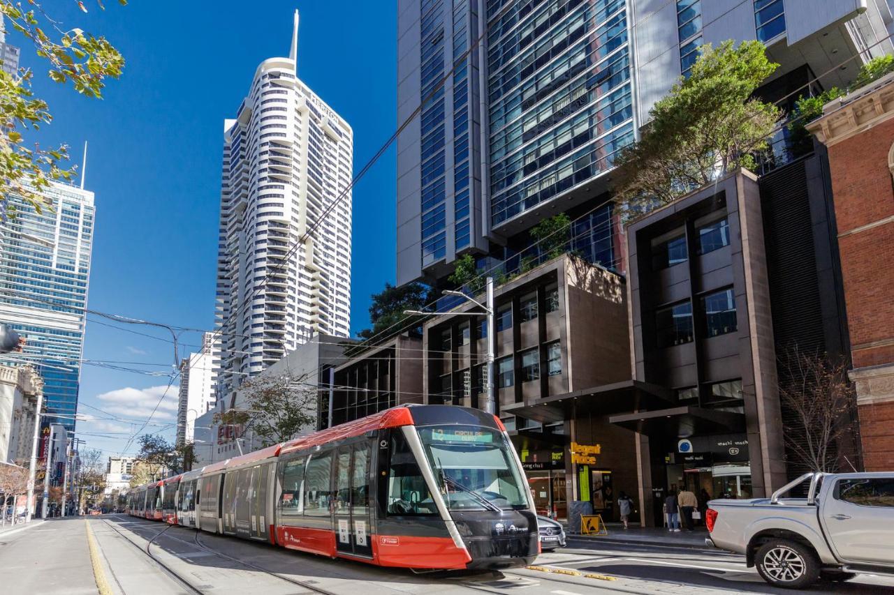 Meriton Suites Kent Street, Σίδνεϊ Εξωτερικό φωτογραφία The photo shows a modern tram or light rail vehicle on a city street. It is positioned in front of a mix of tall buildings, including high-rise apartments and commercial spaces. The setting appears urban and lively, with a clear blue sky and some tre