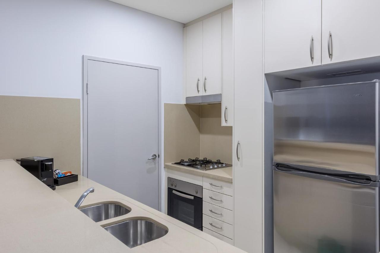 Meriton Suites Kent Street, Σίδνεϊ Εξωτερικό φωτογραφία The photo shows a modern kitchen featuring a sleek design. It includes a light-colored countertop with a double sink, a gas stove, and an oven integrated into the cabinetry. There are upper cabinets for storage, and a stainless-steel refrigerator is 