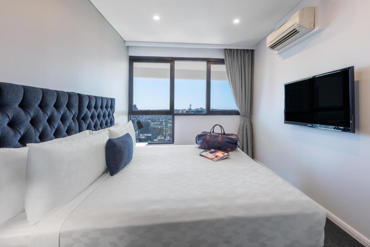 Meriton Suites Kent Street, Σίδνεϊ Εξωτερικό φωτογραφία The photo shows a modern hotel bedroom. It features a large bed with a tufted headboard, dressed in white linens and some decorative pillows. To the side of the bed, there is a stylish bag placed on the bed, along with a magazine or book. The room ha