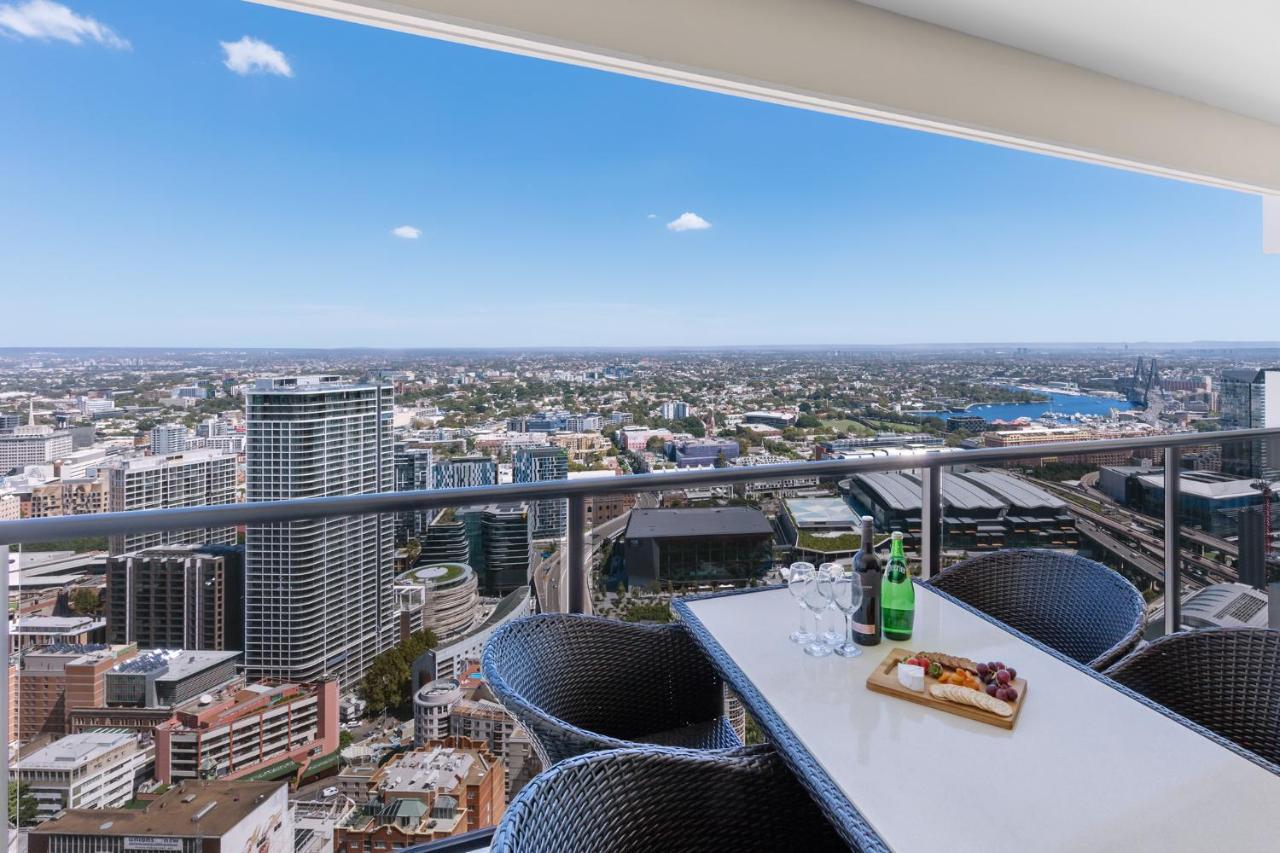 Meriton Suites Kent Street, Σίδνεϊ Εξωτερικό φωτογραφία The photo depicts a rooftop or balcony view of a city skyline from a high elevation. In the foreground, there is a table set with a platter of what appears to be fruits or snacks, along with drinks on the table. The seating around the table consists 