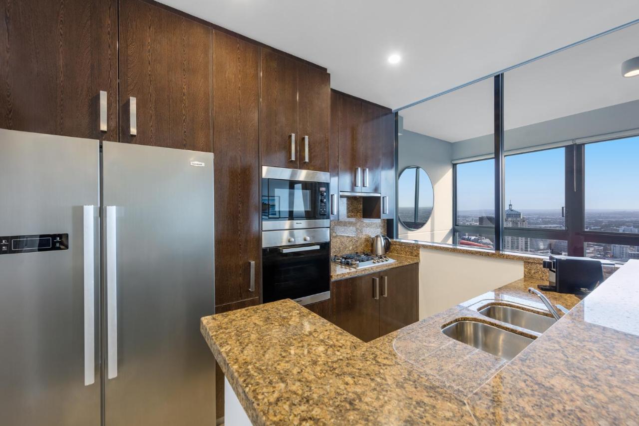 Meriton Suites Kent Street, Σίδνεϊ Εξωτερικό φωτογραφία The photo shows a modern kitchen featuring dark wood cabinetry and a granite countertop. There is a stainless steel refrigerator on the left, alongside built-in appliances including an oven and microwave. A double sink is integrated into the countert