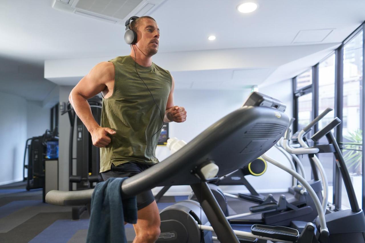 Meriton Suites Kent Street, Σίδνεϊ Εξωτερικό φωτογραφία The image shows a man jogging on a treadmill in a gym. He is wearing a sleeveless green top and headphones. There is a towel draped over the handrail of the treadmill. The gym has large windows, providing natural light, and there are other gym equipm