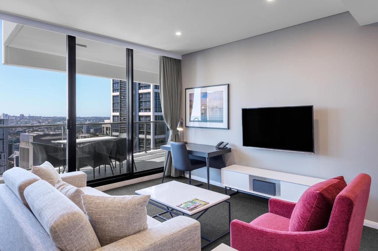 Meriton Suites Kent Street, Σίδνεϊ Εξωτερικό φωτογραφία The photo shows a modern living space with a stylish aesthetic. It features a light-colored sofa and a vibrant pink armchair arranged around a coffee table. A wall-mounted flat-screen TV is set against a light-colored media unit. Large windows allow 