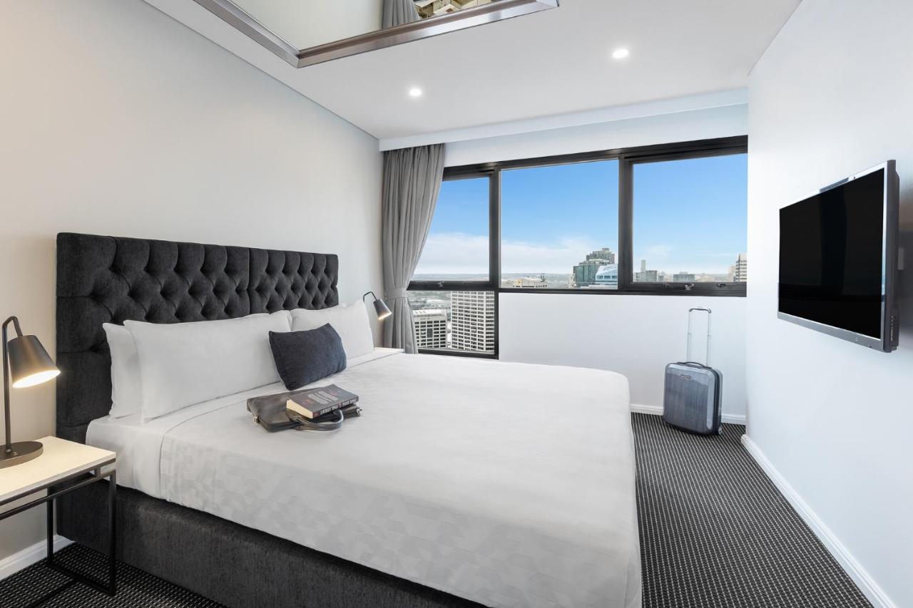 Meriton Suites Kent Street, Σίδνεϊ Εξωτερικό φωτογραφία The photo shows a modern hotel room featuring a large, neatly made bed with a grey upholstered headboard. There is a decorative cushion on the bed, and a small stack of items, possibly books or magazines, placed at the foot of the bed. Next to the be