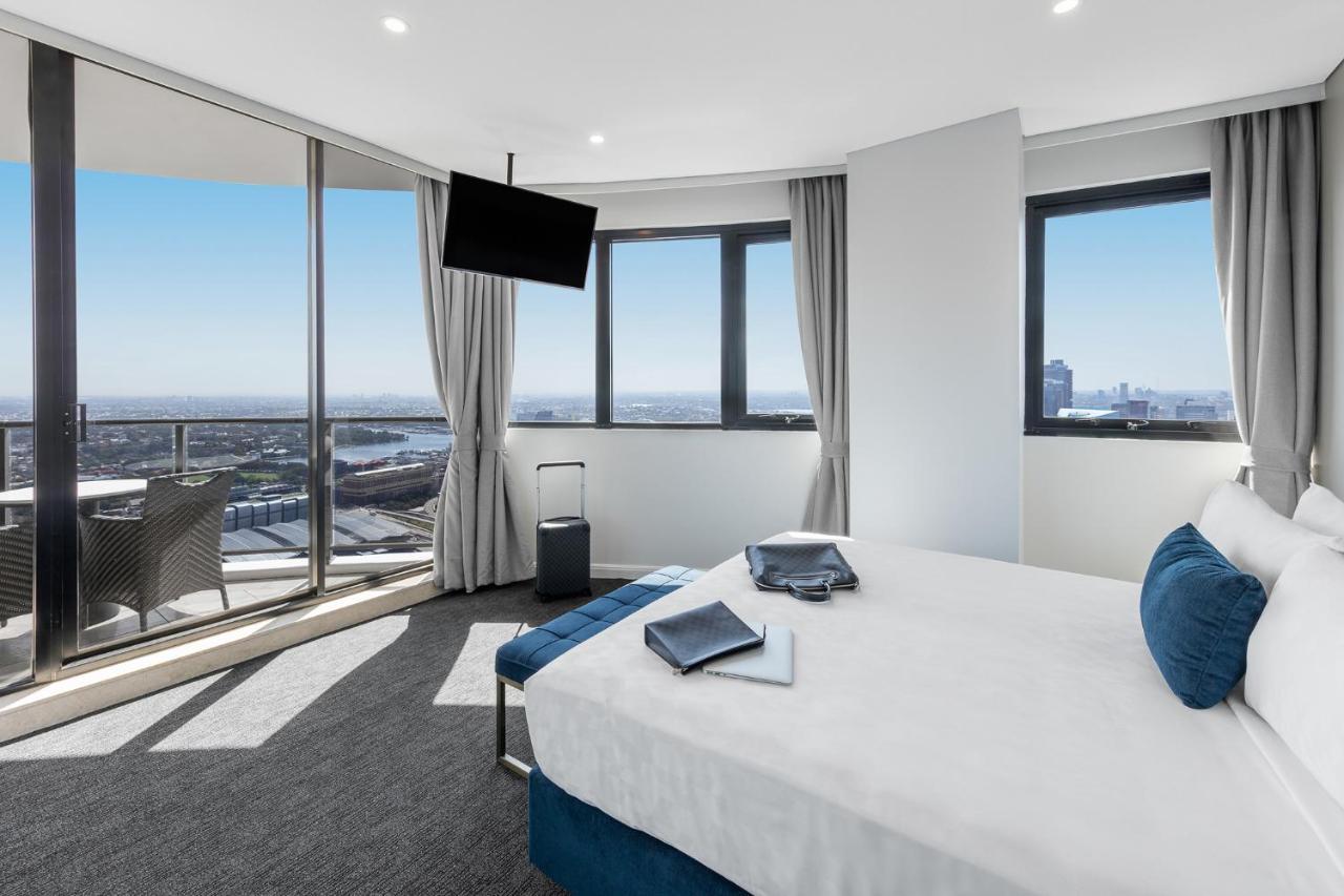 Meriton Suites Kent Street, Σίδνεϊ Εξωτερικό φωτογραφία The photo depicts an elegant hotel room with a modern design. The room features a large bed with white linens and decorative blue pillows. There is a small seating area and a flat-screen TV mounted on the wall. Floor-to-ceiling windows provide a stun