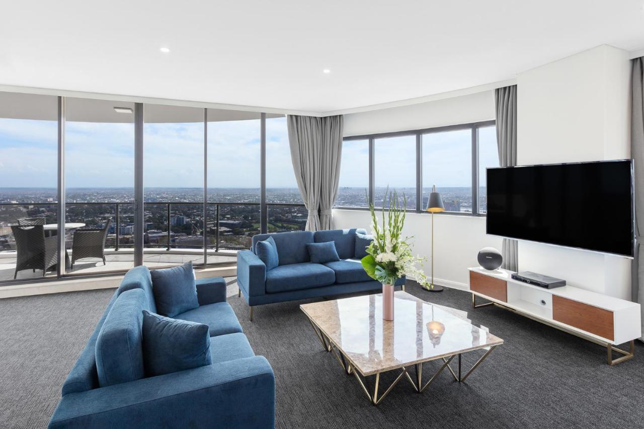 Meriton Suites Kent Street, Σίδνεϊ Εξωτερικό φωτογραφία The photo shows a modern living room with large windows that provide a panoramic view of the city. The room features two blue sofas arranged around a stylish marble-top coffee table. There are decorative items, including a vase with flowers on the ta