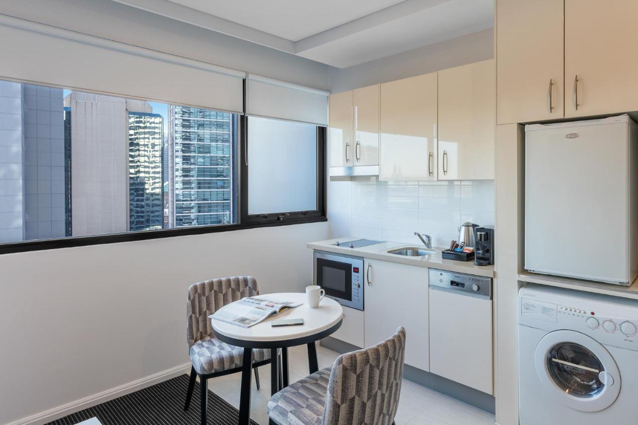 Meriton Suites Kent Street, Σίδνεϊ Εξωτερικό φωτογραφία The photo shows a modern kitchen area featuring light-colored cabinetry. There is a small round table with two patterned chairs nearby. The kitchen includes essential appliances such as a microwave, kettle, and a washing machine, which is positioned 
