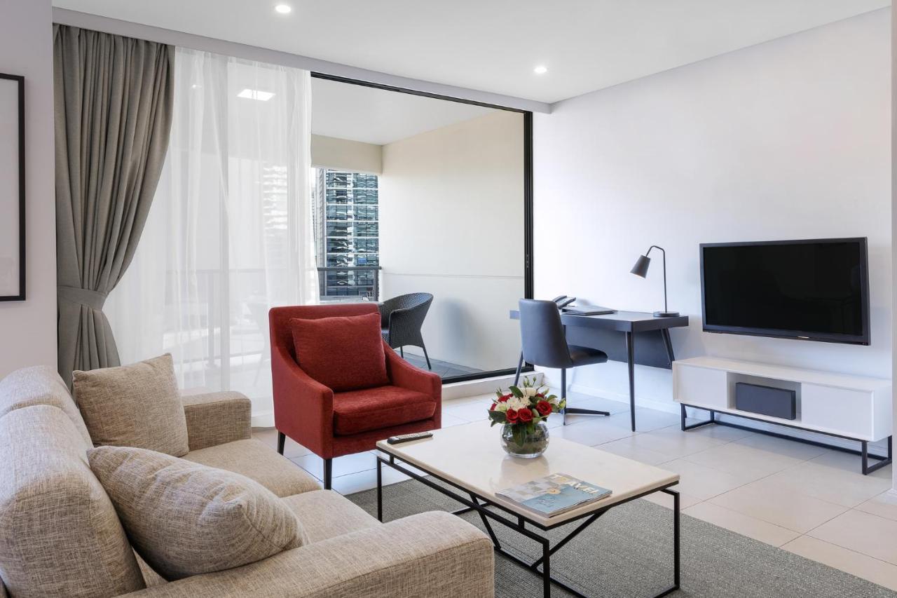 Meriton Suites Kent Street, Σίδνεϊ Εξωτερικό φωτογραφία The photo shows a modern living space featuring a comfortable cream-colored sofa and a vibrant red armchair. A coffee table sits in front of the sofa, adorned with a small vase of flowers and a magazine. There is a television mounted on the wall oppo