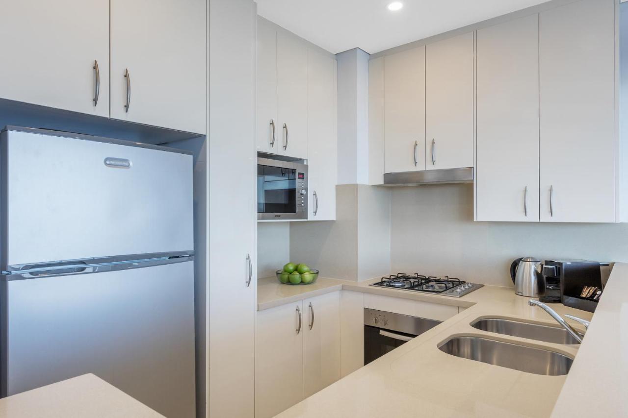 Meriton Suites Kent Street, Σίδνεϊ Εξωτερικό φωτογραφία The image shows a modern kitchen with a sleek design. It features light-colored cabinetry, a stainless steel refrigerator, and a built-in microwave. There is a gas stove with an overhead range hood and a double sink with a fashionable countertop. A b