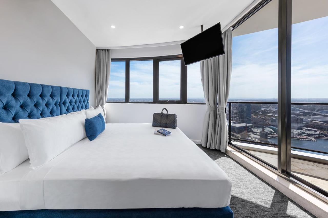 Meriton Suites Kent Street, Σίδνεϊ Εξωτερικό φωτογραφία The photo shows a modern hotel room featuring a large bed with white linens and a tufted blue headboard. There is a blue decorative pillow on the bed. In the background, large windows are letting in natural light and offering a view of the outside. A
