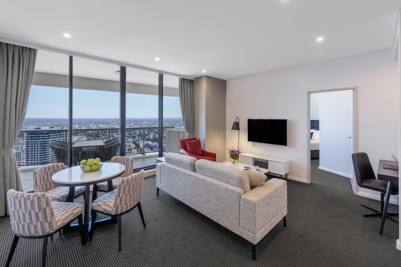 Meriton Suites Kent Street, Σίδνεϊ Εξωτερικό φωτογραφία The photo shows a modern and stylish living area in a high-rise apartment. It features large windows that offer a view of the city. The space includes a comfortable gray sofa, a circular dining table with four chairs, and a red accent chair. A televi